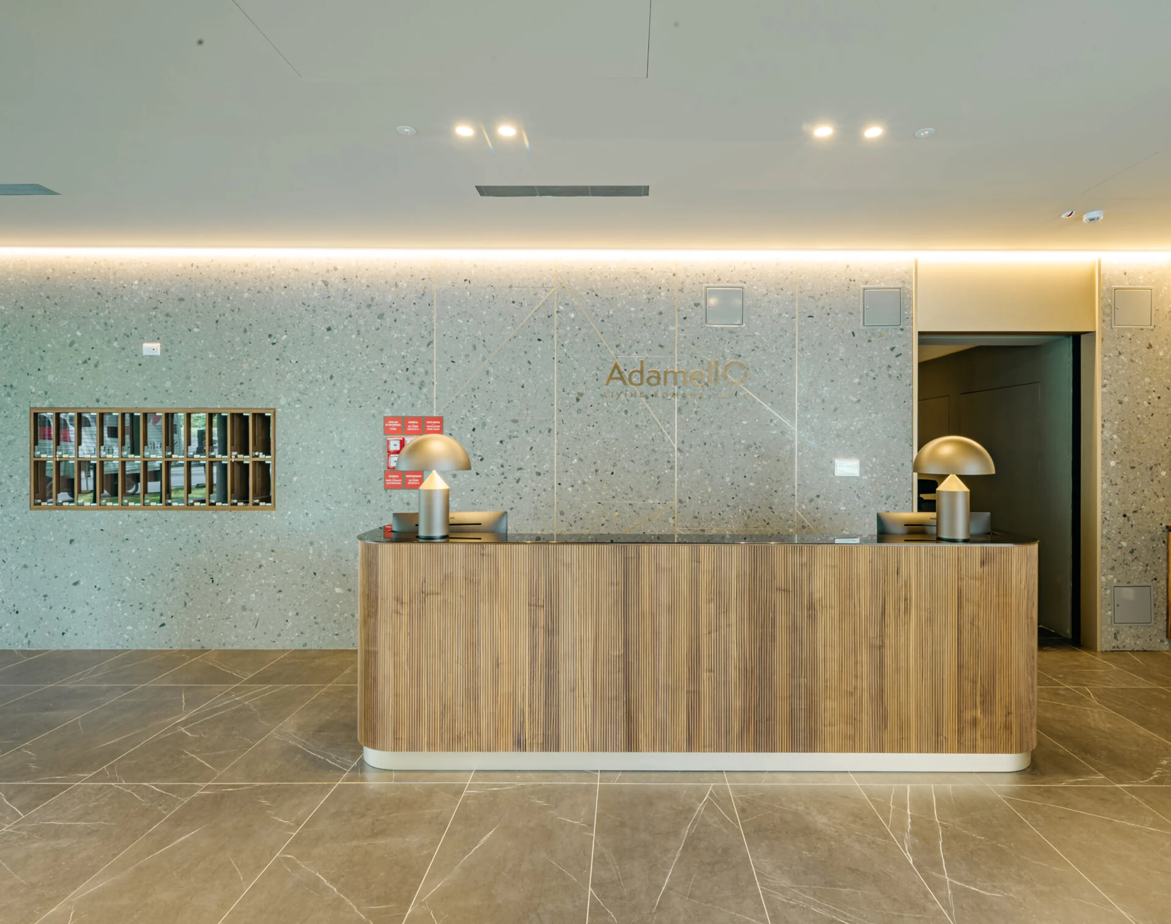 Hall d'ingresso del complesso Adamello Living Romana Yards: reception in legno con piano scuro, parete a casellario in Agglotech SB130 Aggloceppo levigato con inserti in ottone e pavimento in marmo - posa in opera Trapattoni Marmi
