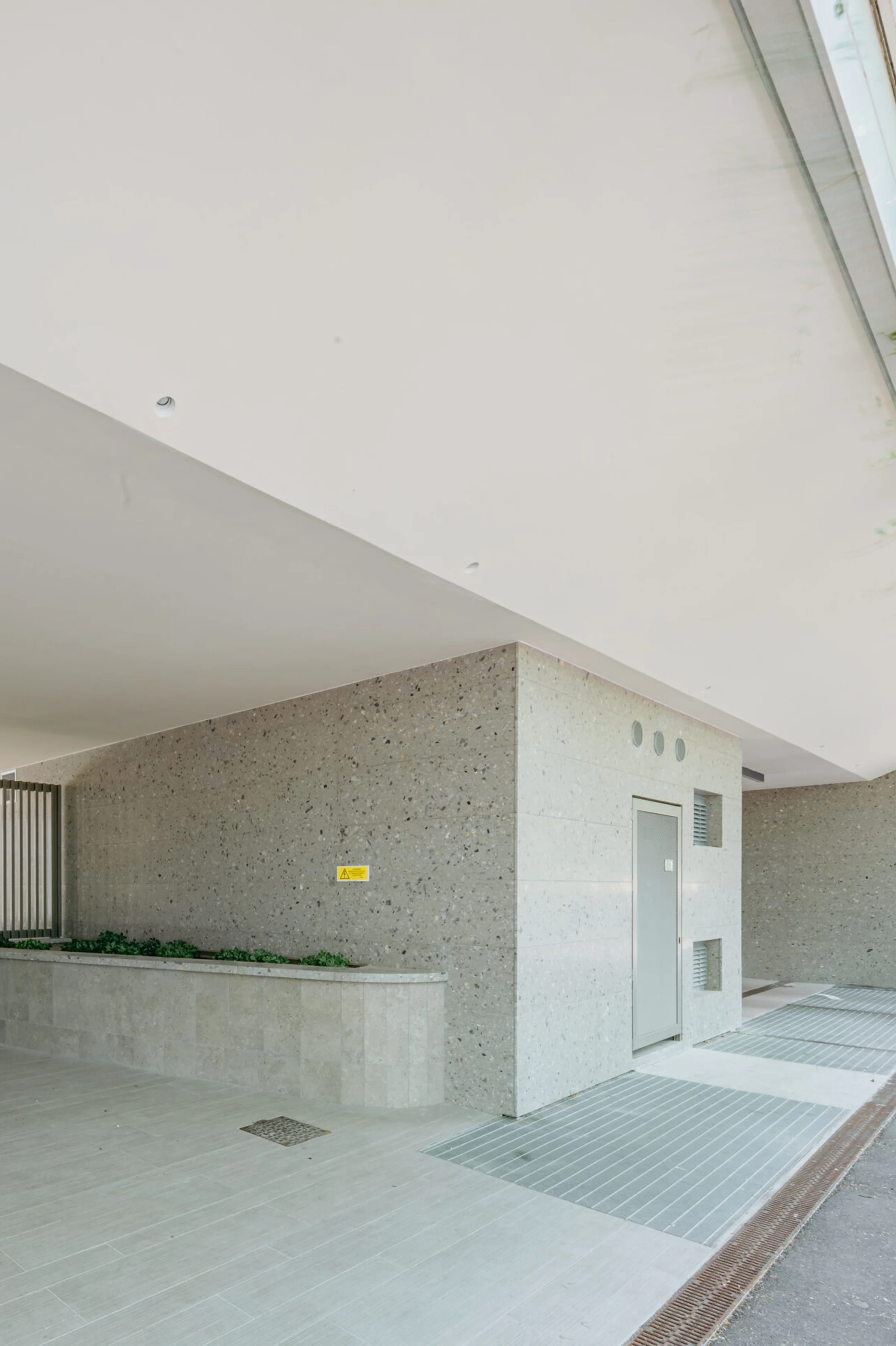 Dettaglio del portico esterno della Torre Adamello con zoccolatura e rivestimento a parete in Agglotech SB130 Aggloceppo levigato, pavimentazione in pietra chiara e corpi illuminanti a soffitto - Trapattoni Marmi, Milano