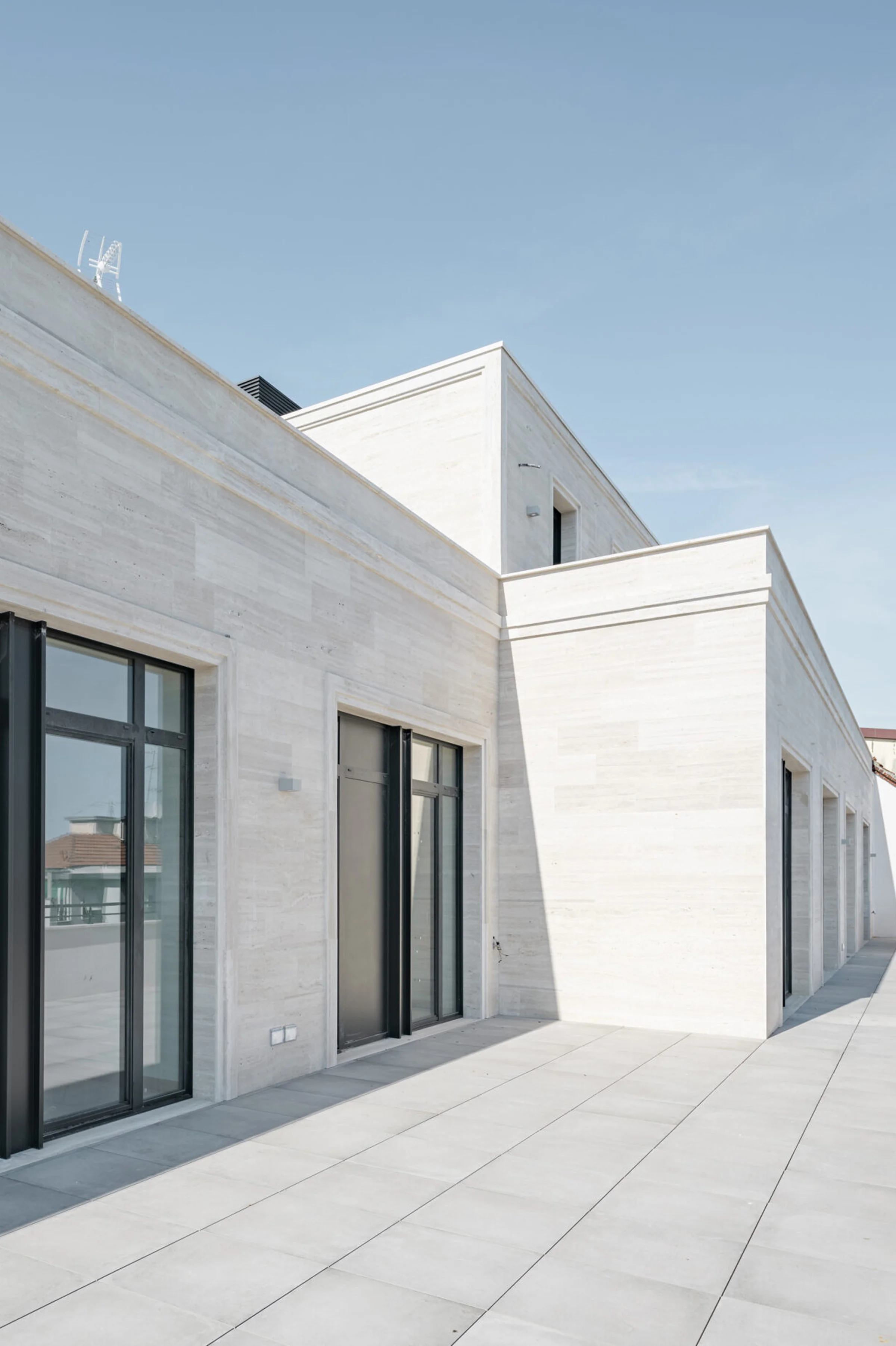 Terrazza del Washington Building a Milano con pareti e volumi della penthouse rivestiti in Travertino Romano a venatura orizzontale, infissi neri e pavimentazione in pietra chiara - fornitura e posa in opera Trapattoni Marmi